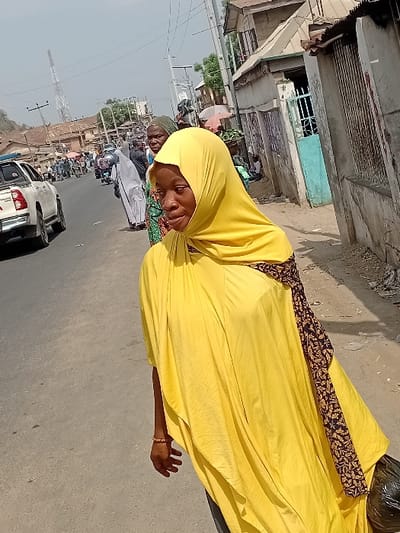 A woman in a yellow hijab and dress walks alongside a busy street. The dress has a dark brown and gold pattern on the upper back. Behind her, a line of cars moves along the road. Buildings line the left side of the frame, with power lines overhead. Other people are also visible in the background.