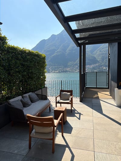 A sunny day is depicted on a balcony overlooking a lake and mountains in Pognana Lario, Italy. The balcony is furnished with a modern sofa and two wooden chairs with light-colored cushions, and a small wooden table between them. A black metal and glass canopy extends over part of the balcony. A tall green hedge lines the left side of the balcony, and a black metal railing separates the balcony from the view. The lake, with its surface sparkling in the sun, is visible beyond the railing, as are lush green mountains in the distance. A dark wall is on the right side of the balcony, with a white planter containing an umbrella. The floor is tiled with large, light-colored square tiles.