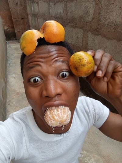 A medium close-up, high-angle shot depicts a young male subject with dark skin, short dark hair, and a white, textured short-sleeved t-shirt, performing a playful pose against a rough-textured, light brown concrete block wall in Aramoko-Ekiti, Nigeria. The subject's eyes are wide open, eyebrows raised creating horizontal forehead wrinkles, and mouth open. A peeled orange segment cluster is positioned inside the subject's mouth, with visible pulp/juice at the lower lip. Two orange peels, appearing to be from separate oranges, are balanced on top of the subject's head, resembling ears or horns. The peel on the right side of the head (viewer's left) is more intact, while the other appears partially broken. The subject holds an unpeeled, whole orange with an orange-yellow skin and dark spots in his right hand, positioned near the left side of his face. The foreground shows a light grey concrete surface.