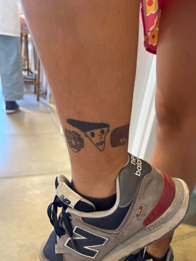 An angled view shows a person's lower leg with three cartoon tattoos: a slice of pizza with a face, a blob character, and a circular pizza. The leg is fair-skinned and slightly hairy. The ankle is adorned with a gray and navy New Balance 574 sneaker. Visible in the background are a tan-colored floor, a patterned garment, and a blurred room setting with furniture.