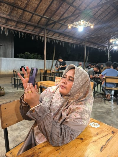 The image depicts a woman wearing a patterned headscarf and long-sleeved blouse, seated at a wooden table in what appears to be an outdoor dining area. She is holding a purple smartphone and appears to be taking a photo or video. The dining area has a thatched roof supported by wooden beams and columns, and it is illuminated by hanging light fixtures. Other people are seated at tables in the background, some eating and others conversing. The environment suggests the location may be in Cempeudak, Indonesia.
