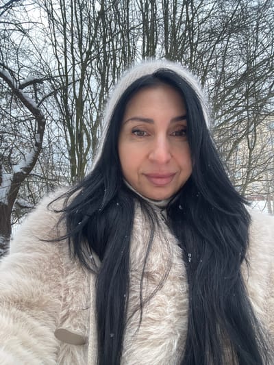 The image depicts an adult female outdoors in a snowy, wooded environment within the city of Syasstroy, Russia. The woman, positioned centrally and looking directly at the camera, has long, dark hair, a medium skin tone, and a subtle smile. She is dressed in a light cream or beige long-haired faux fur coat, exhibiting a visible light-colored toggle closure on the left side, over what appears to be a light-colored turtleneck. A fuzzy, light-colored headband or hat portion is visible, framing the top of her dark hair. Small, distinct snowflakes are scattered on her hair and coat. The background features numerous bare deciduous trees, their branches lightly dusted with snow, under an overcast sky. A light-colored building is faintly discernible through the trees in the upper right background. The scene conveys a cold, winter setting with active or recent light snowfall.