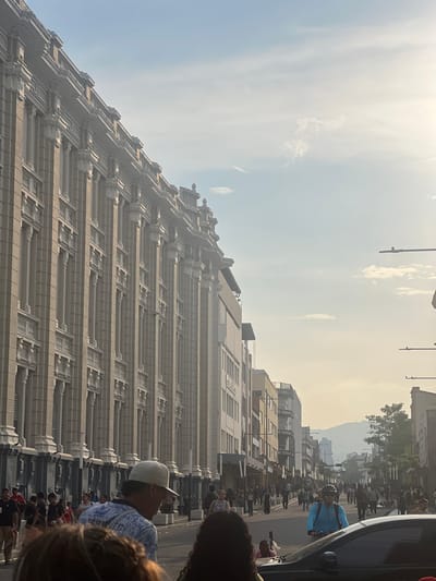 The image shows a street scene in San Salvador, El Salvador, featuring the heavily ornamented facade of the Teatro Nacional de El Salvador. The tall, light-colored building dominates the left side of the frame and is constructed with repetitive vertical and horizontal elements. A crowd of people is walking along the street in the foreground and background, with some figures partially obscured by a car on the right. The sky is bright with a haze, and distant buildings and a mountain are visible in the background.