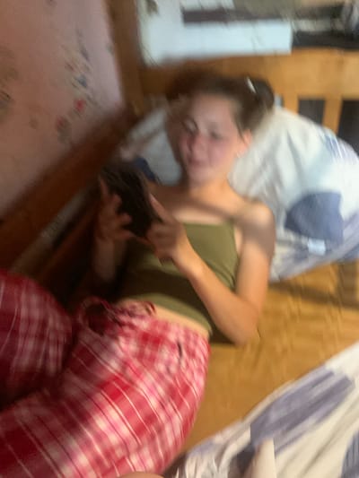 In Hoholeve, Ukraine, a young girl is lying on a bed, holding a dark-colored smartphone in her hands. She is wearing a green tank top and red and white plaid pajama bottoms. The bed is covered with a brown-colored sheet and a blue and white striped blanket. The background shows a wooden bed frame and a wall with a floral pattern. The image is slightly blurred, suggesting movement or a low-resolution camera.