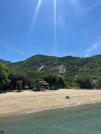 This media file captures a scenic coastal view in Hoa Thang, Vietnam. The scene is dominated by a lush, green mountain covered in dense vegetation that rises dramatically from the sandy shore. The mountain's steep, rocky face is partially visible through the foliage.
In the foreground, a golden sand beach stretches along the edge of the tranquil, light green ocean. Gentle waves with white foam are lapping at the shore. A single, dark-colored jet ski is parked on the sand near the water's edge. Further up the beach, several other objects covered by tarps are visible, possibly boats or equipment. A small cluster of buildings with reddish-brown roofs is nestled at the base of the mountain, surrounded by trees and vegetation. Some parked vehicles are visible near these buildings.
The sky is a clear, bright blue with a few wispy white clouds. A strong lens flare extends diagonally across the left side of the frame, indicating bright sunlight. The time of day appears to be midday, with the sun high in the sky. The weather is clear and sunny, contributing to the idyllic and peaceful atmosphere of the location. The overall impression is one of a tropical paradise, inviting relaxation and outdoor activities. No visible text is present in the media file.