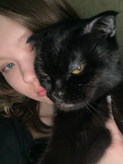 A black cat with golden eyes rests its head on a person's face. The cat's black fur is visible, highlighting its head and part of its body. The person, presumably a young female, is looking toward the camera. A silver necklace can be seen around her neck. The background is dark and undefined, drawing focus to the interaction between the cat and the person.