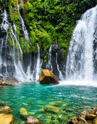 The image depicts a powerful waterfall cascading down a lush green cliff face into a clear turquoise pool. The water is a vibrant blue-green, with visible rocks and pebbles beneath the surface. A large, rough-textured boulder sits in the middle of the pool, partially submerged by the falling water. The cliff is densely covered in verdant vegetation, with water seeping and flowing down in numerous rivulets alongside the main waterfall. The overall environment suggests a tropical rainforest setting. There are no people visible in the image. The scene appears to be a natural landscape, likely a tourist attraction or a secluded natural wonder.