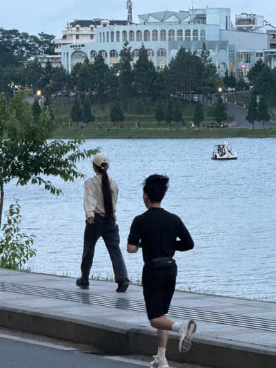 The image depicts a lakeside scene in Xuan Huong Lake, Da Lat, Vietnam. In the foreground, two individuals are engaged in physical activity. A person in the foreground is running away from the camera, wearing a black long-sleeved shirt, black shorts, white socks, and white sneakers. They have a fanny pack around their waist. To the left, another individual is walking away from the camera, wearing a light-colored long-sleeved shirt, dark pants, and a light-colored cap. Their long dark hair is visible down their back.
The background features a body of water, presumably Xuan Huong Lake, with a small pedal boat carrying at least three people in the distance. Beyond the lake, a tree-lined area leads up to a collection of buildings. One prominent building is light blue with arched windows and appears to be a large structure, possibly a hotel or a public building. Other buildings with a mix of architectural styles, including white facades and dark roofs, are visible further in the background, interspersed with trees. The overall lighting suggests it is either early morning or late evening. The ground in the immediate foreground is a paved walkway with a textured surface.