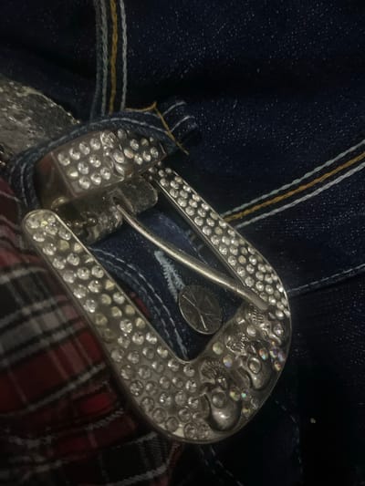 The image shows a close-up of a belt buckle and denim fabric. The belt buckle is ornate, made of silver-toned metal and encrusted with numerous clear rhinestones. It features a skull motif with multiple skulls incorporated into its design, also decorated with rhinestones. A metal prong extends from the buckle, ready to be inserted into a belt hole. The buckle is positioned over dark blue denim, which appears to be part of a pair of jeans. The denim has visible stitching, including some yellow and white threads, characteristic of high-quality denim construction. A small, circular metal object, possibly a coin or part of the belt, is visible beneath the prong. To the right of the buckle, a plaid fabric, primarily red and black, is visible, suggesting it is part of the same garment or an accessory worn with it. The lighting is dim, with reflections on the rhinestones and metal, indicating an indoor setting. There is no text visible in the image. The focus is on the decorative elements of the belt buckle and the texture of the fabrics.
