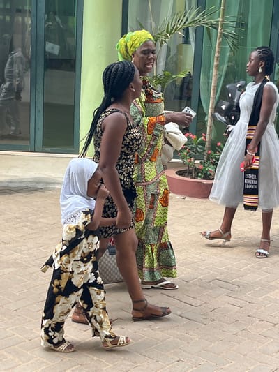 The scene depicts four individuals outdoors on a paved area, with an implied location in the city of Madina, Ghana. The ground consists of light brown rectangular pavers. In the background, a light green building with large glass doors reflects a partially visible figure in light clothing. Potted plants with green foliage and small red flowers, along with slender palm-like trees, are situated along the building's base.
From left to right:
A young child, possibly female, wears a white hijab with lace trim and a black, white, and gold floral patterned jumpsuit with light-colored sandals. This child is holding the left hand of an adult female standing next to her.
The adult female, positioned centrally, has braided dark hair and wears a sleeveless black and gold/brown leopard-print patterned dress, reaching above the knee. She wears brown flat sandals and carries a beige and brown patterned handbag (possibly Gucci print) on her left forearm.
Adjacent to her is another adult female with a bright green, yellow, and orange patterned headscarf. She wears a long dress with similar green, orange, and white patterns. Her mouth is open, indicative of speaking or singing. She holds a white clutch bag in her left hand and a silver smartphone in her right. Her footwear consists of white flip-flops.
To the far right stands a young female wearing a white sleeveless dress with a tulle-like skirt. Over the dress, she wears a black and patterned graduation stole with the text "ESTHER KUMUWAA BROWN" and horizontally striped patterns in red, gold, and black. She holds a cluster of black balloons in her left hand. Her footwear consists of silver open-toed sandals with clear heels.