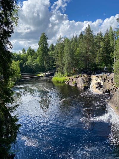 A wide, dark blue river flows in the foreground, with white foam swirling on its surface. The river is bordered by rocky banks and lush green trees on both sides. In the middle ground, a small waterfall cascades over dark rocks. A wooden bridge spans the river above the waterfall, and several people are visible on the bridge and on the rocks near the waterfall. The background is filled with a dense forest of tall green trees. The sky above is blue with scattered white clouds. The scene depicts a natural park setting, likely in Ruskeala, Russia, with the river and waterfall being prominent features.