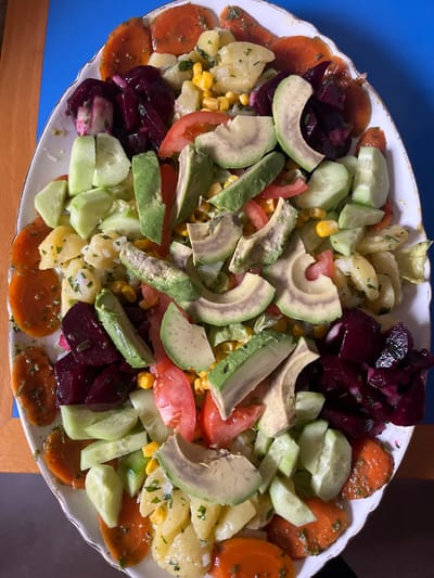 The scene, located in Le Puy-en-Velay, France, presents an overhead view of a large, oval-shaped serving platter filled with a mixed salad. The platter is white with a scalloped rim and a thin, light-colored edge detail. The salad components are distributed throughout the platter and include slices of green avocado, chunks of dark red beetroot, pale green cucumber pieces, and sliced red tomatoes. A central base or filler consists of cubed potatoes mixed with bright yellow corn kernels and chopped green herbs, consistent with parsley. Marinated or cooked orange carrot slices, also garnished with green herbs, are arranged along the perimeter of the platter. The platter rests on a surface that is partly a light brown wooden table and partly a vibrant blue surface visible to the left and top of the platter.