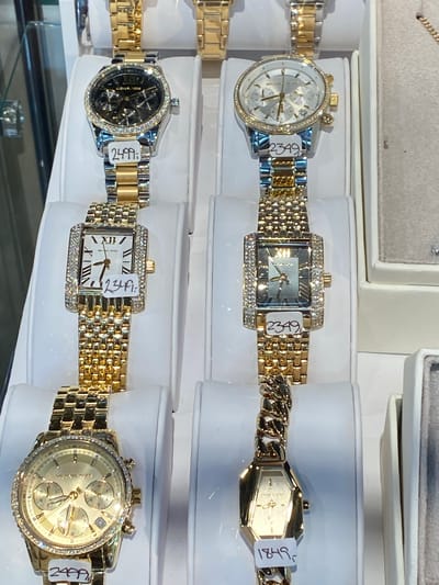 The image displays a close-up view of several gold-colored watches displayed on white stands. The watches vary in design, with some featuring diamond-encrusted bezels and others having more elaborate faces with multiple dials. Price tags are visible on some of the watches, indicating prices such as "2499,-", "2349,-", and "1849,-". The background is blurred, suggesting a retail environment such as a jewelry store. The watches are the primary focus, presented in a well-lit manner to highlight their details and craftsmanship. The overall impression is one of a high-end product display.