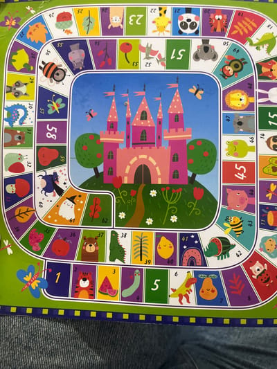 The image is a close-up, top-down view of a colorful children's board game, likely a fantasy or animal-themed game, laid out on a textured surface that appears to be denim jeans. The board is circular in shape, with numbered spaces featuring illustrations of animals, fruits, a castle, and magical creatures like a ghost and a unicorn. The game path winds its way around a central illustration of a pink castle. The dominant colors are green, purple, blue, and various bright hues used for the illustrations. There are no people visible in the image, and no recognizable landmarks or details that would suggest a specific location within the city of Gorodets, Russia. The scene is static and depicts a game board.