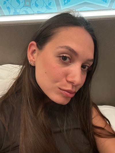 A woman with long, dark brown hair is reclining against a dark headboard and a white pillow. Her face, angled towards the camera, features smooth skin, light pink lips, brown eyes, and delicate eyelashes. She has multiple piercings in her ear. The background includes a window with a patterned design on the left. The overall impression is relaxed and natural.
