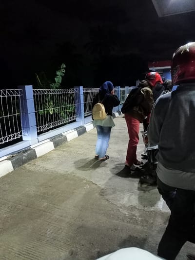 The image captures an outdoor scene at night in Cimaung, Indonesia, featuring several people and motorcycles under artificial light. In the foreground, a woman wearing a blue hijab, black top, and light blue jeans stands with her back partially to the camera, carrying a small beige backpack. She is facing towards a blue and white metal fence with a wire mesh pattern, beyond which dark foliage and trees are visible against the night sky.

To her right, a person in a brown jacket and red pants is positioned next to a motorcycle, appearing to tend to it or stand by it. Further to the right and closer to the foreground, another person is seen from behind, wearing a grey shirt and dark pants, with a red helmet on their head. Another red helmet is partially visible on the far right, suggesting at least two motorcycles are present.

The ground is concrete pavement, marked by shadows cast by the subjects and an adjacent black and white striped curb. The overall lighting suggests a brightly lit area against a dark night, possibly a roadside stop or a gathering point. There is no visible text in the image. The atmosphere appears calm, indicative of a casual stop or waiting period.