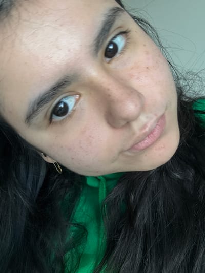 A close-up, angled selfie shows a person with dark hair and fair skin. The subject is wearing a green top and a gold safety pin is visible near their left ear. The background is a plain white wall. There is no contextual information in the image to determine a location in Austin, United States of America, or any other specific location.