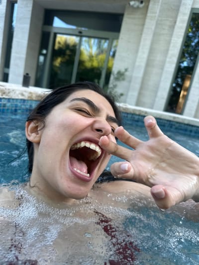 The image shows a young woman with dark hair submerged in a pool, with bubbles rising around her. She is smiling broadly, with her mouth open and her eyes closed. Her right hand is raised, with her fingers spread apart. The pool is surrounded by a tiled edge, and in the background, a modern building with large glass doors and windows is visible. The overall scene suggests a leisurely moment spent in a pool on a warm day. The location is specified as Dzoraghbyur, Armenia.