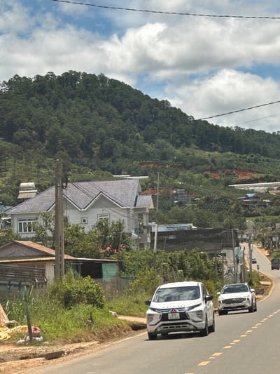 The image depicts a bright, daytime scene on a road in Thôn 1, Vietnam. A white Mitsubishi Xpander is in the foreground, driving on the right side of the paved road. A white Hyundai Santa Fe is behind it, also on the right side of the road. The road curves to the right, leading uphill. On the left side of the road, there is a grassy embankment with some vegetation and a large, light-colored house with a gray tiled roof. Further up the hill, more houses and buildings are visible, interspersed with dense green trees. The background is dominated by a heavily forested mountain slope under a partially cloudy sky. The overall impression is a rural or semi-rural residential area in a hilly region.