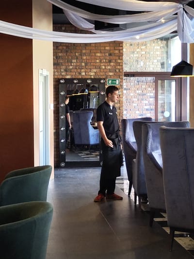 A young male server stands in a restaurant, facing right, wearing a black polo shirt, black apron, black pants, and red sneakers. He is positioned in front of a large, illuminated mirror that reflects a blue armchair and a checkered floor pattern. The restaurant features exposed brick walls, hanging white fabric decorations, and dark upholstery on the chairs. The scene is captured from a medium shot, with the server as the primary subject. The location is identified as Zhukovsky, Russia.
