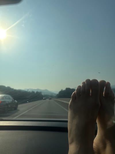 This is a first-person perspective, eye-level shot taken from inside a vehicle traveling on a highway. The foreground is dominated by a pair of bare feet resting on the car's dashboard, with the toes facing the camera. The feet appear to be illuminated by the bright sun. Through the windshield, the highway stretches into the distance, with a line of cars visible on the road. The scenery on either side of the highway consists of rolling hills covered in trees, and the sky is a hazy blue with the sun prominently featured in the upper left corner, casting lens flare. The image appears to be taken during daylight hours, likely in the late morning or early afternoon. The overall impression is one of a leisurely road trip. The location is identified as a highway in Namyangju-si, South Korea.