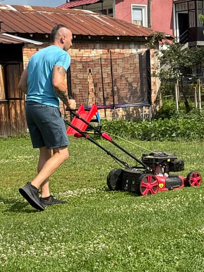 An adult male with a mohawk haircut and tattoos on his arm is mowing a grassy yard with a red and black lawnmower. He is wearing a blue t-shirt and dark shorts. The yard is covered in green grass and small white flowers. In the background, there are buildings with red roofs and brick walls. The scene appears to be a residential area in Oreshak, Bulgaria, on a sunny day.