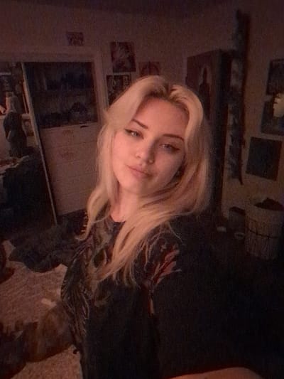 The image displays a young female subject indoors. The precise geographical location of Area D (Elaho/Garibaldi), Canada, cannot be visually determined from the provided image; the setting is an enclosed room. The subject, positioned centrally, has shoulder-length, wavy, light blonde hair and light-colored eyes, with a small stud piercing on her right nostril and visible winged eyeliner. She is wearing a dark-colored top featuring a faint, multi-toned pattern on the shoulders and upper arms, and looks directly at the camera with a neutral expression, her head slightly tilted.
The background, dimly lit with warm light and exhibiting a grainy quality, shows a room with light-colored walls. To the subject's left, a door frame or opening reveals a light-colored dresser or cabinet. Posters are affixed to the wall above this area, and a reflective surface, possibly a mirror, contains a blurred reflection of a figure. To the subject's right, a tall, dark furniture piece is visible alongside additional posters or framed artworks. A woven basket or hamper is partially visible on the floor in the far right. The lower left foreground shows a textured light brown or beige rug, with an indistinct, dark shape present on its surface.