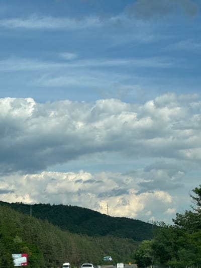 The image captures a scenic view of a mountainous landscape under a dramatic sky, with a highway passing through. The foreground features a road with several vehicles, including white vans and cars, indicating traffic. To the left, a billboard advertising "ALPIN" is visible, partially obscured by trees.
The terrain rises steeply on the left, covered in dense green coniferous forest. A power line tower is situated on the crest of the hill, with more such towers visible further in the distance. The sky is a prominent feature, filled with layers of cumulus clouds, ranging from bright white to dark gray, creating a dynamic and somewhat overcast atmosphere. The upper portion of the sky is a lighter blue with wispy cirrus clouds. The overall impression is a natural landscape with human infrastructure intersecting it, likely on the outskirts of Rajana, Bulgaria, given the context.