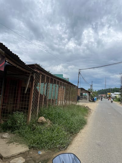 The image depicts a roadside scene in Rantau Pauh, Indonesia, under a cloudy sky. In the foreground to the left, a derelict building with a corrugated metal roof and a rusted metal fence is visible. Overgrown grass and some debris, including plastic bags and a rolled-up mat, surround the building. A concrete path is also present. The road extends into the distance on the right, with several motorcycles and a few people riding them. Power lines and poles are strung across the scene. Further down the road, more buildings, trees, and a distant communication tower can be observed. The overall impression is of a rural or semi-urban area with signs of aging infrastructure.