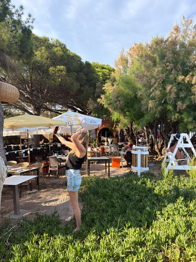 A woman is taking a photograph with her phone at the beach restaurant "Royal Bliss" in Mont-roig del Camp, Spain. She is wearing a black top and denim shorts. Tables and chairs are visible under sun umbrellas. Behind the restaurant are trees and dense green bushes. A small white structure is on the right.