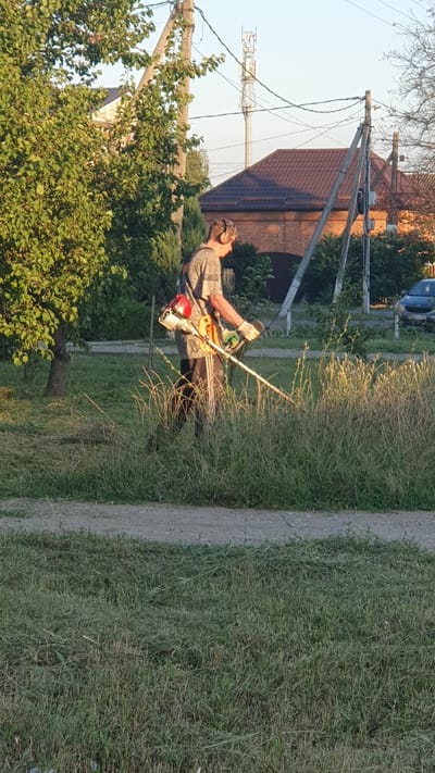 In Ust-Labinsk, Russia, a person is seen trimming a grassy area with a string trimmer. The individual, wearing a patterned shirt, dark pants with a white stripe, gloves, and headphones, is positioned on a slightly elevated patch of land adjacent to a paved surface. Behind them, a tree with lush green foliage is visible, as are several buildings with brown roofs and brickwork. A tall cell tower stands in the distance, alongside utility poles and overhead wires. The lighting suggests it is either early morning or late afternoon, with the sun casting long shadows and highlighting the dry grass. A dark-colored car is parked on the right side of the frame.