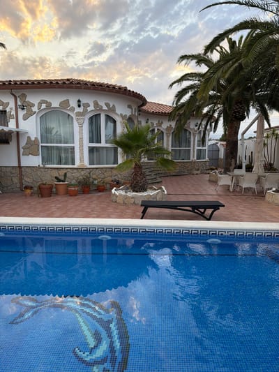 A patio area with a swimming pool is in the foreground. A house with white walls and arched windows is in the background. Palm trees are visible on the right side of the house. The sky is cloudy.