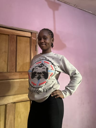 The scene depicts a young female indoors in Agbogugu, Nigeria. She is positioned centrally, facing the camera with a slight smile, revealing dimples on both cheeks. Her hair is dark and styled in braids. She wears a grey long-sleeved sweatshirt featuring a prominent circular graphic on the chest. This graphic includes a black, stylized dog-like face within a compass-like design with red and blue accents, and the text "FASHION EVISU" below. Her left hand rests on her hip. She is also wearing a form-fitting black skirt or trousers. The background consists of a solid pink wall to the right and a light-colored wooden door with vertical panels to the left. A dark, elongated shadow is cast on the upper right portion of the pink wall, extending towards the ceiling.