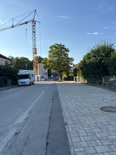 The image depicts a street scene in Höhenkirchen-Siegertsbrunn, Germany, on a clear, sunny day. A large construction crane dominates the left side of the frame, its tall yellow and gray structure reaching towards the blue sky. In the foreground, a white camper van is parked on the asphalt road, with "Dethleffs" and "Advant-Age" visible on its side. Behind the camper, a blue truck is also parked, along with portable toilets and stacks of concrete blocks, indicating ongoing construction work. The right side of the road features a paved sidewalk made of gray rectangular stones, bordered by a curb and a small patch of grass. Trees with lush green foliage line the street, casting shadows on the sidewalk. In the background, houses with tiled roofs are visible, suggesting a residential or mixed-use area. A manhole cover is present on the sidewalk. The overall impression is one of a typical day in a suburban German town with construction underway.