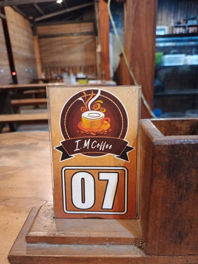 A close-up shot focuses on a table number sign, marked '07', for 'I. M Coffee' in Geulumpangmeujim-Jim, Indonesia. The sign features a brown and yellow coffee cup logo. It is placed on a worn wooden surface next to a wooden container, likely a utensil or condiment holder. In the blurred background, one can discern a wooden structure, potentially a part of the cafe's interior or a surrounding building. Seating is visible in the background.