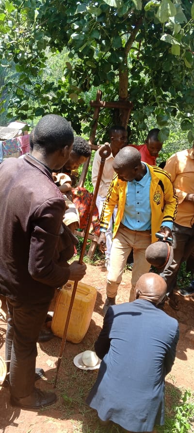 A group of people, mostly men, are gathered outdoors under a tree. One man in a brown suit holds a cane and a yellow plastic container. A man in a yellow jacket with a blue shirt looks down at the container. Another man in a blue suit is squatting near a hat. Other individuals are standing around, observing. The setting appears to be rural, with trees and foliage visible in the background.