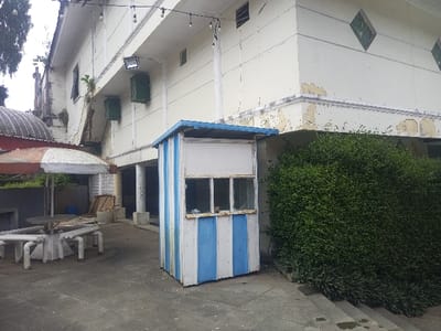 The image depicts an outdoor area adjacent to a multi-story building in Cipanas, Indonesia. In the foreground, a small, weathered kiosk or booth stands on the right, featuring vertical blue and white stripes, a blue roof, and two front windows with dirty panes. To the left, a white outdoor table with attached circular benches, constructed from cylindrical components, surrounds a collapsed, faded red and white striped umbrella. The ground is paved with dark grey square tiles.
The background is dominated by an off-white building exhibiting significant peeling paint and discoloration, particularly along its cornices and ledges. The upper floor features decorative diamond-shaped elements and small rectangular windows. Below a horizontal projection, multiple green rectangular units, possibly air conditioning components or vents, are mounted on the wall, along with an exterior floodlight. String lights are suspended above the building's facade. A dense green hedge grows along the base of the building on the right, rising from a tiered concrete planter. Further left, a corrugated metal roof is partially visible, and a more dilapidated structure with a tree branch extends upwards. No individuals are present in the scene.