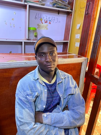 The image shows a young Black man wearing a blue denim jacket over a blue patterned t-shirt and a beige baseball cap, seated with his arms crossed. He is looking directly at the camera with a neutral expression. The background suggests an indoor setting, possibly a shop or stall, with shelves visible above and a wooden counter in front of him. The shelves contain various items, including a clear plastic container with a green lid. There are some drawings and handwritten numbers on the wall behind him, reading "6140786730". The overall lighting is warm and appears to be artificial. The location is identified as Barakin, Nigeria.