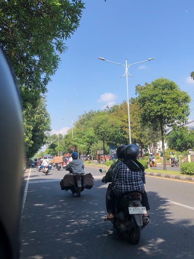 The image captures a traffic scene on a sunny day in Banjarkemantren, Indonesia. The perspective is from a motorcycle, looking forward down a two-lane road lined with trees. Several other motorcycles are visible, some carrying passengers and luggage. In the foreground, a motorcycle with two occupants, one wearing a black and white checkered shirt and black hijab, is the primary subject. The motorcycle has a license plate reading "W 4725 WS 07-30". Further down the road, other motorcycles are in motion, and a few cars are present in the background. The environment appears to be a suburban or semi-urban area with lush green trees lining the street and a clear blue sky overhead. Streetlights are visible along the road. The overall impression is a typical scene of daily commuting in this Indonesian locale.