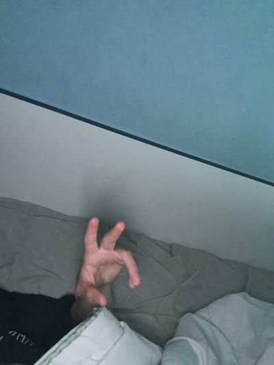 The image captures an intimate indoor scene, likely a bedroom in Priseltsi, Bulgaria. The main subject is a human hand, pale in complexion, with fingers splayed open as if stretching or relaxed. It emerges from beneath light grey bedding, resting on a textured grey sheet or blanket that covers the bed.

A significant visual element is a sharp, diagonal line that divides the left side of the frame into two distinct panels. The leftmost section is a solid, muted blue, while the section immediately to its right and adjacent to the bed is a lighter, almost off-white or very light grey. These panels appear to be part of a wall or a large piece of furniture, such as a headboard or closet.

In the upper right and lower right portions of the frame, a fluffy, light-colored (possibly white or very pale green) duvet or comforter is visible, partially covering a person implied to be in the bed. A small piece of black fabric with faint, illegible white text is visible just below the duvet in the bottom right corner; the characters are indistinct but could resemble "mn" or other fragmented letters. The overall lighting is soft and diffused, suggesting a quiet moment indoors, possibly during the early morning or evening hours. There are no direct cues about the weather or specific emotions, but the setting implies rest or transition.