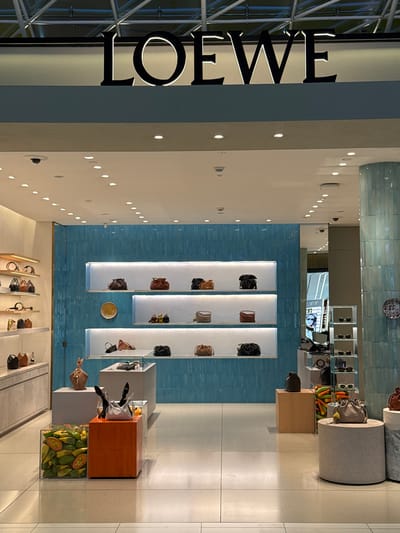 The image depicts the interior of a luxury retail store, likely a handbag or fashion boutique, in Nong Prue Subdistrict, Thailand. The store features modern, minimalist displays with white shelving and light-colored walls. A prominent "LOEWE" logo is visible on the left side of the image, indicating the brand.
Various handbags are displayed on shelves. One display features a clear cube filled with sliced fruits, possibly as a decorative element. Another display shows a cylindrical concrete pedestal with a handbag on top. The store lighting is soft and ambient, suggesting it could be evening or the store is designed with low lighting. The overall aesthetic is clean, sophisticated, and upscale. No people are visible in the image, and the scene appears to be a quiet moment within the store. The tiled blue wall in the background adds a touch of color and texture to the otherwise neutral palette.