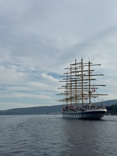A large, multi-masted sailing ship, identified as the "Royal Clipper," is captured from a side-rear perspective, sailing on a body of water. The ship has a white and blue hull, with numerous masts and rigging supporting sails, though the sails themselves are not deployed. Several people are visible on the deck of the ship. In the background, a verdant, hilly coastline with scattered buildings is visible, suggesting a temperate climate. The sky is overcast with scattered clouds. The scene is likely set in the Bay of Kotor, near the village of Lepetane, Montenegro, given the characteristic landscape. The water is dark blue and appears moderately choppy.
