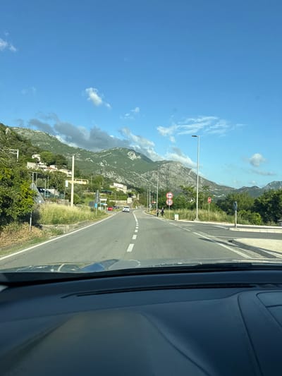 The image depicts a road in Kaluđerac, Montenegro, as seen from inside a car. The road is asphalt with white lane markings. Signage indicates a speed limit of 50 km/h and a stop sign. Mountains are visible in the background under a blue sky with scattered clouds. Vegetation lines the sides of the road. There's a passing car ahead on the road.