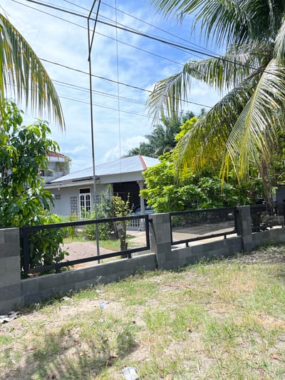 The image depicts a residential property in the city of Langsa, Indonesia. A grey, single-story house with a metal roof is visible, featuring white-framed windows and doors. The house is surrounded by lush green foliage, including trees and bushes, with palm fronds hanging from the right side of the frame. A dark metal fence with a grey concrete base encloses the property. The ground in front of the fence is covered in dry grass and dirt, with some scattered debris. The sky above is blue with scattered white clouds. Electrical wires are suspended across the upper portion of the image.