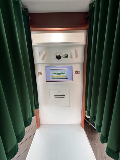 The image depicts an ATM-like machine in what appears to be an indoor setting. The machine has a screen displaying the words "Sukhalakela Mart". There are green curtains partially obscuring the top and bottom of the machine. The environment includes a white wall and wood-paneled accents. Based on the text on the machine, it is located in Candikuning, Indonesia.