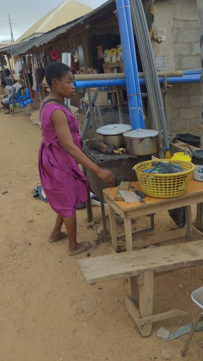 A woman in a purple dress stands near a cooking area on a sandy ground in what appears to be Masaka, Nigeria. She faces towards the right of the frame, towards several pots sitting atop a charcoal grill. The pots are covered with lids and presumably cooking food. Beside the pots is a yellow plastic basket. The cooking area appears to be near a building with construction supplies, including blue plastic pipes. A partially visible poster is affixed to the building. A wooden structure is in the foreground on the right. The sky is overcast.