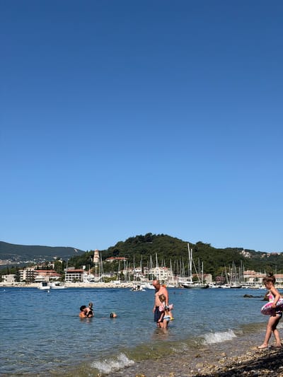 The image depicts a sunny day at a coastal town, identified as Meljine, Montenegro. The foreground shows a rocky beach with clear, shallow water lapping at the shore. Several people are visible in the water and on the beach. In the water, a group of three individuals, likely a mother and two children, are swimming. To the right, a woman with a pink inflatable ring walks along the shore. Further into the water, a man stands with two young girls, one of whom is wearing arm floaties. In the background, a picturesque bay is filled with numerous sailboats moored in a marina. Behind the marina, buildings line the waterfront, and a lush green hillside rises, topped with a church spire and more buildings scattered amongst the trees. The sky is a clear, vibrant blue.