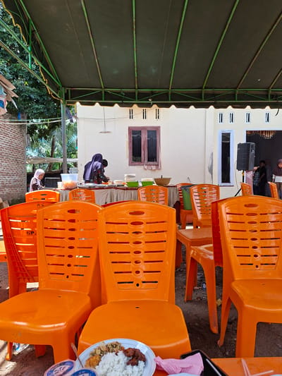 The image depicts an outdoor seating area set up for an event, likely a meal or gathering, under a green tarp canopy supported by a metal frame. The ground is dirt and gravel. In the foreground, several bright orange plastic chairs are visible, arranged in rows. One chair has the partial inscription "GP" on its backrest. On a table in the lower right corner, a plate of food is present, containing white rice and various side dishes, alongside two small blue plastic containers and a pink napkin.
Further back, a table is set up with plates, bowls, and food containers, and several people are interacting around it. Two individuals, appearing to be women wearing headscarves, are attending to the food. Another person, possibly a child, is also near the food table. To the right, a black speaker is positioned on a stand near an open doorway. The building behind the tables has cream-colored walls and multiple windows. Trees and a brick wall are visible in the background to the left. The scene appears to be in a semi-rural or residential area, potentially in Pie, Indonesia, given the architectural style and general atmosphere.