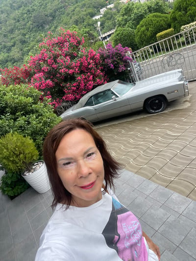 A woman with brown hair and red lipstick is taking a selfie outdoors. She is wearing a white t-shirt with a colorful graphic on it. To her left, a silver classic car is parked on a paved driveway. Behind the car, a lush green hillside covered with trees is visible. In the foreground, there are vibrant pink flowering bushes and manicured green shrubs. A white planter with small stones and some greenery sits to the woman's right. The scene appears to be set in a residential area with a fence and decorative metalwork in the background. The lighting suggests it is daytime, possibly a bright but overcast day due to the soft shadows.