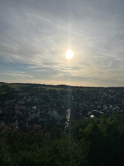 An elevated, high-angle outdoor shot captures a panoramic view of a small town or village, identified as Untergruppenbach, Germany, situated within a valley or depression surrounded by undulating hills and agricultural land. The sun is prominently featured high in the upper-right quadrant of the sky, emitting strong glare and lens flares, suggesting either late afternoon or early morning.

The sky exhibits a gradient of light blue to pale grey, with wispy cirrus or altocumulus cloud formations. Two distinct contrails are visible: one traversing horizontally across the upper sky, and another thin, vertical contrail positioned directly below the sun.

In the midground, the town of Untergruppenbach unfolds, characterized by a dense cluster of residential buildings with predominantly dark-tiled, pitched roofs and light-colored facades. A prominent white church with a tall, pointed spire and a clock tower is visible towards the left side of the town cluster. Streets with a few visible vehicles wind through the settlement. The sun's rays illuminate the rooftops and upper sections of buildings on the right side of the town, creating bright highlights, while areas in shadow appear darker.

Beyond the town, the landscape transitions into a mosaic of cultivated fields displaying various shades of green and brown, interspersed with dark green forested areas on the surrounding hills. The horizon line is relatively flat, with distant land features subtly visible.

The immediate foreground consists of a dense canopy of dark green deciduous trees, heavily shadowed, indicating the elevated vantage point from which the photograph was taken. No individuals are discernible within the frame.
